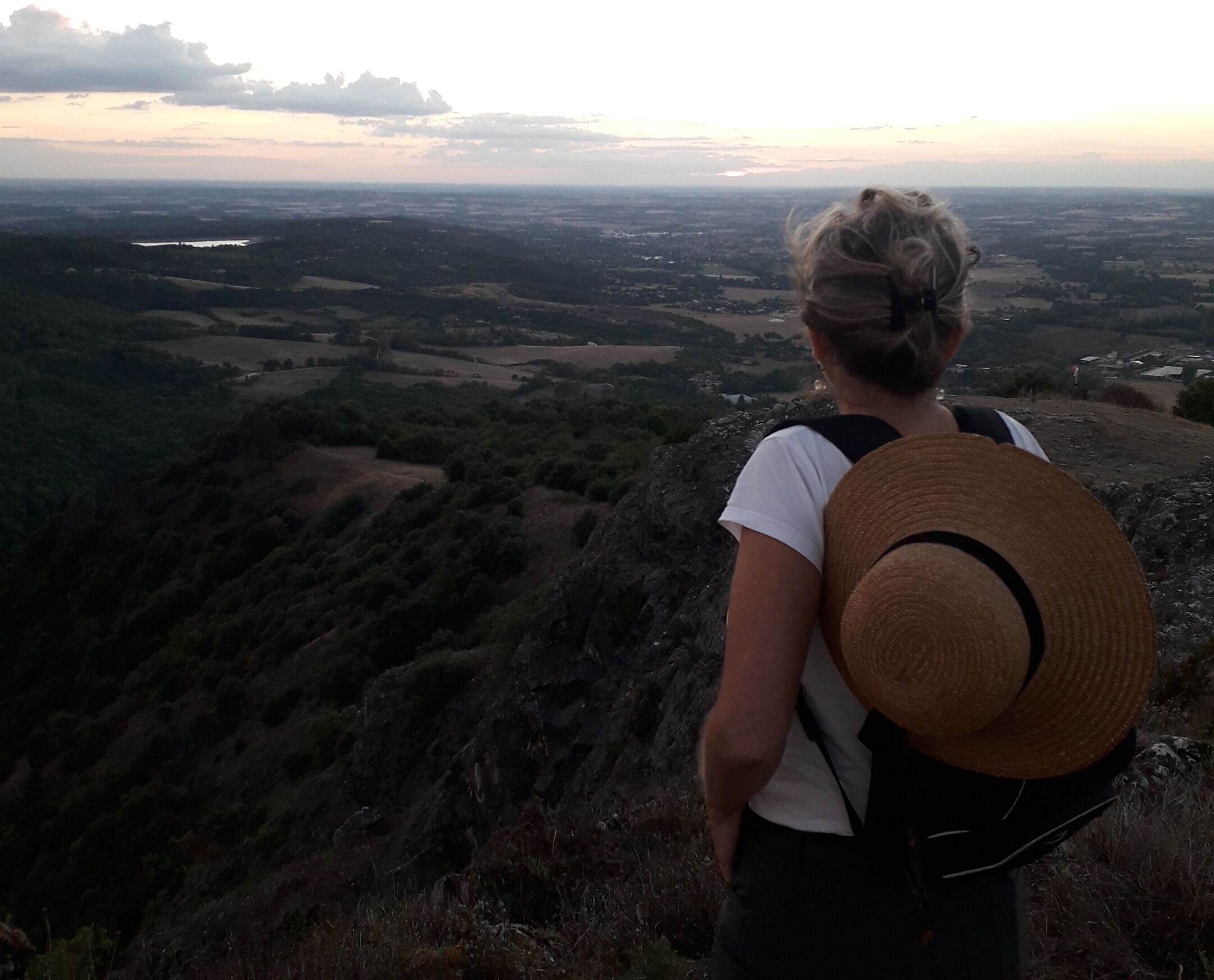 A person with a straw hat and a backpack overlooking a valley with fields and a distant body of water.