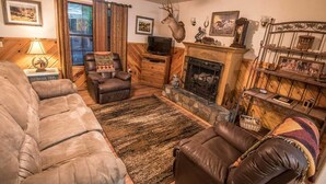 A cozy living room with a fireplace, leather sofas, a television, and a deer head mounted on the wall. - Julia Cabin is a quaint cabin that sleeps up to 6 people in the existing bedding. (BROKEN BOW)