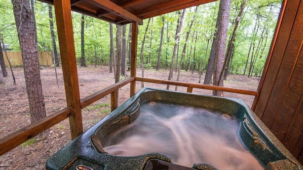 A hot tub in a wooden structure surrounded by trees. - Julia Cabin is a quaint cabin that sleeps up to 6 people in the existing bedding. (BROKEN BOW)