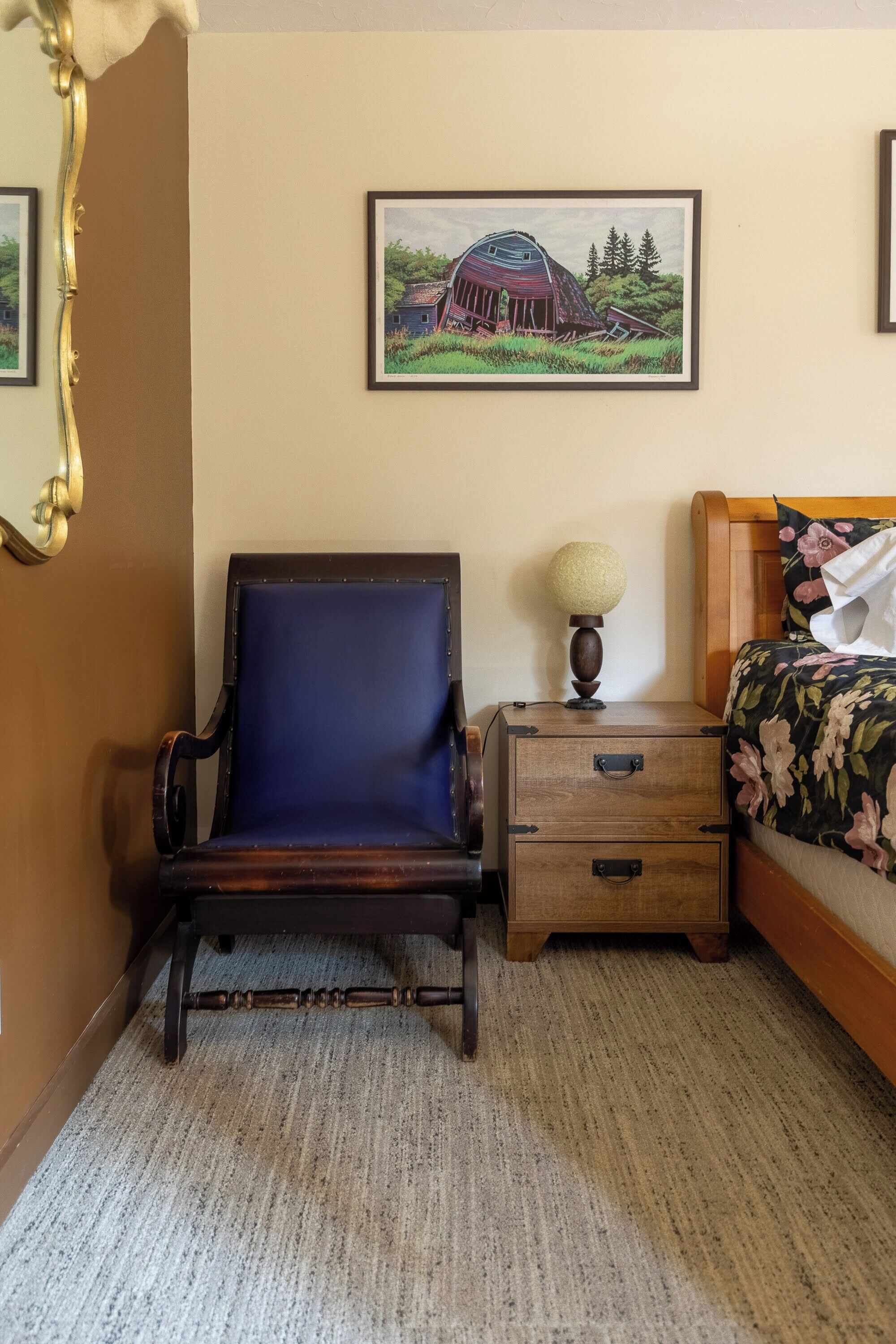 A room with a blue upholstered chair, a wooden nightstand with a lamp, a bed with floral bedding, and a framed picture of a barn on the wall.