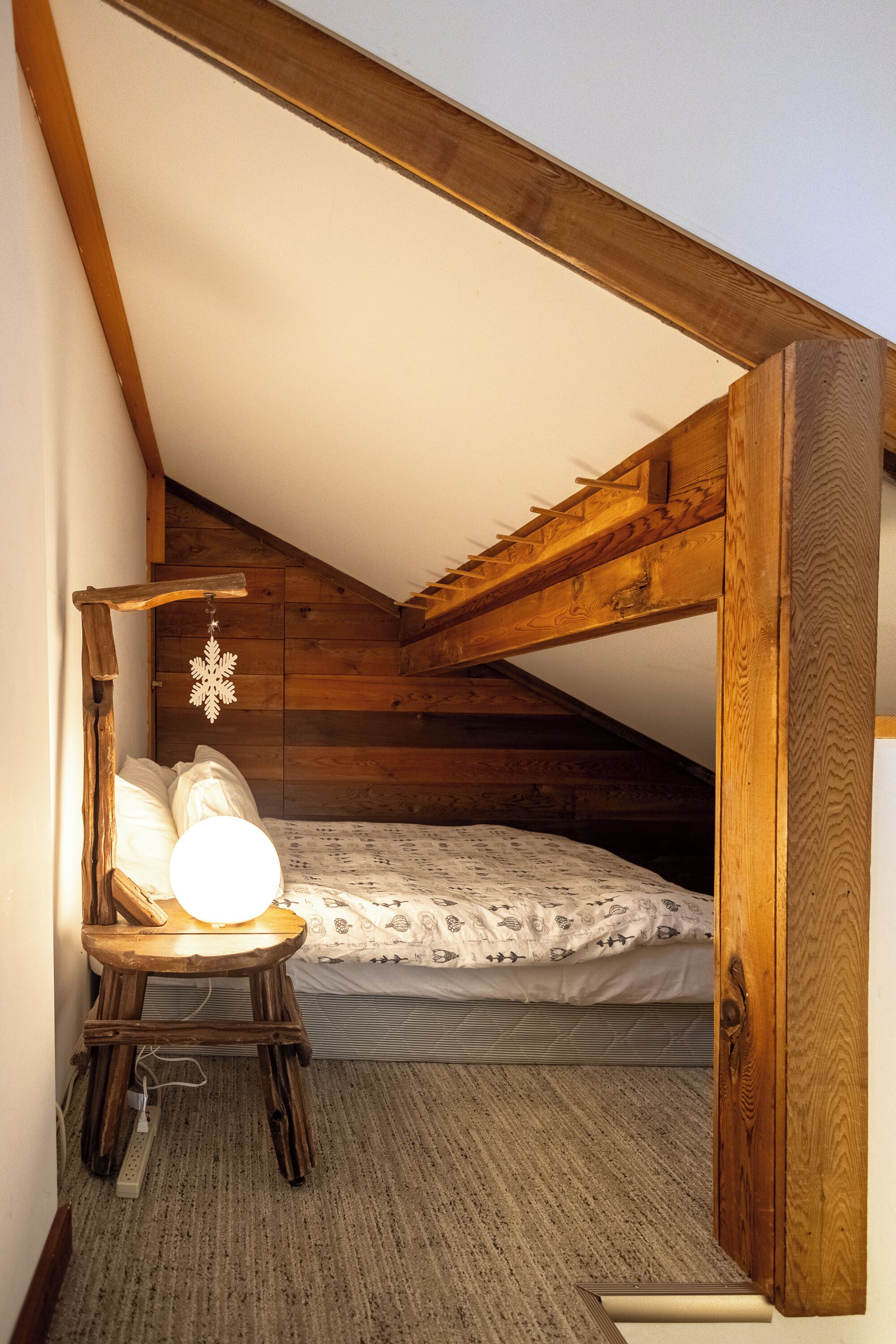 Un pequeño dormitorio en el ático con techo inclinado, estructura de cama de madera, mesita de noche con lámpara y una decoración de copo de nieve colgante.