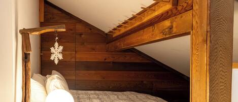 Un pequeño dormitorio en el ático con techo inclinado, estructura de cama de madera, mesita de noche con lámpara y una decoración de copo de nieve colgante.