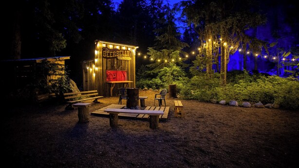 A rustic wooden structure labeled "The B&B Saloon" with a red neon sign, surrounded by a natural setting with benches and string lights. - Rams Head Inn (Rossland)