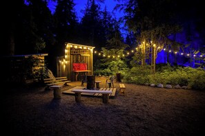 A rustic wooden structure labeled "The B&B Saloon" with a red neon sign, surrounded by a natural setting with benches and string lights. - Rams Head Inn (Rossland)