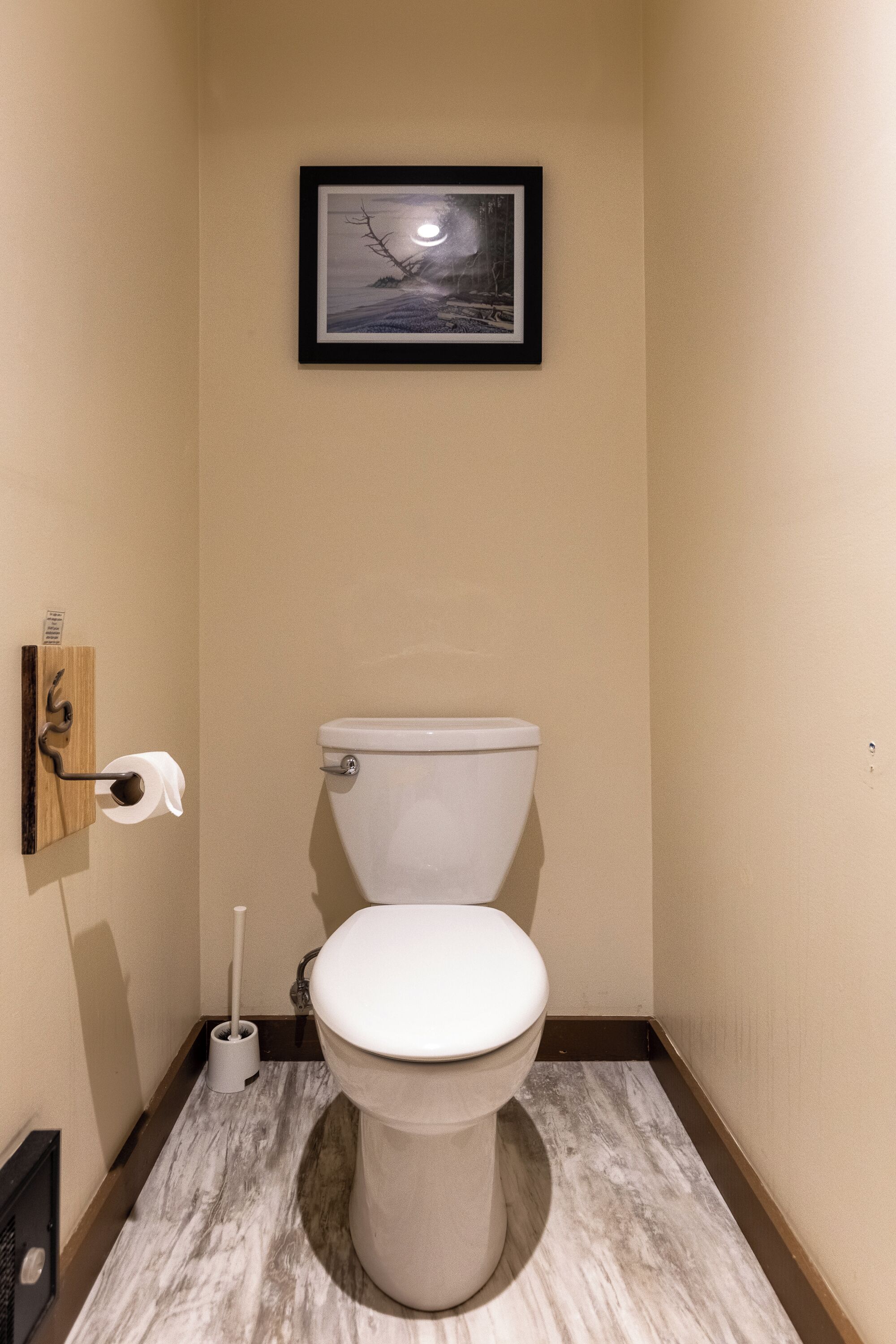 Une petite salle de bain étroite, avec des toilettes, un tableau encadré au mur et un porte-rouleau de papier toilette.
