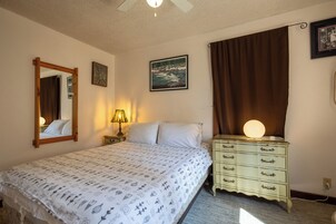 Un dormitorio con cama, mesita de noche, cómoda, espejo, un cuadro en la pared y ventilador de techo.