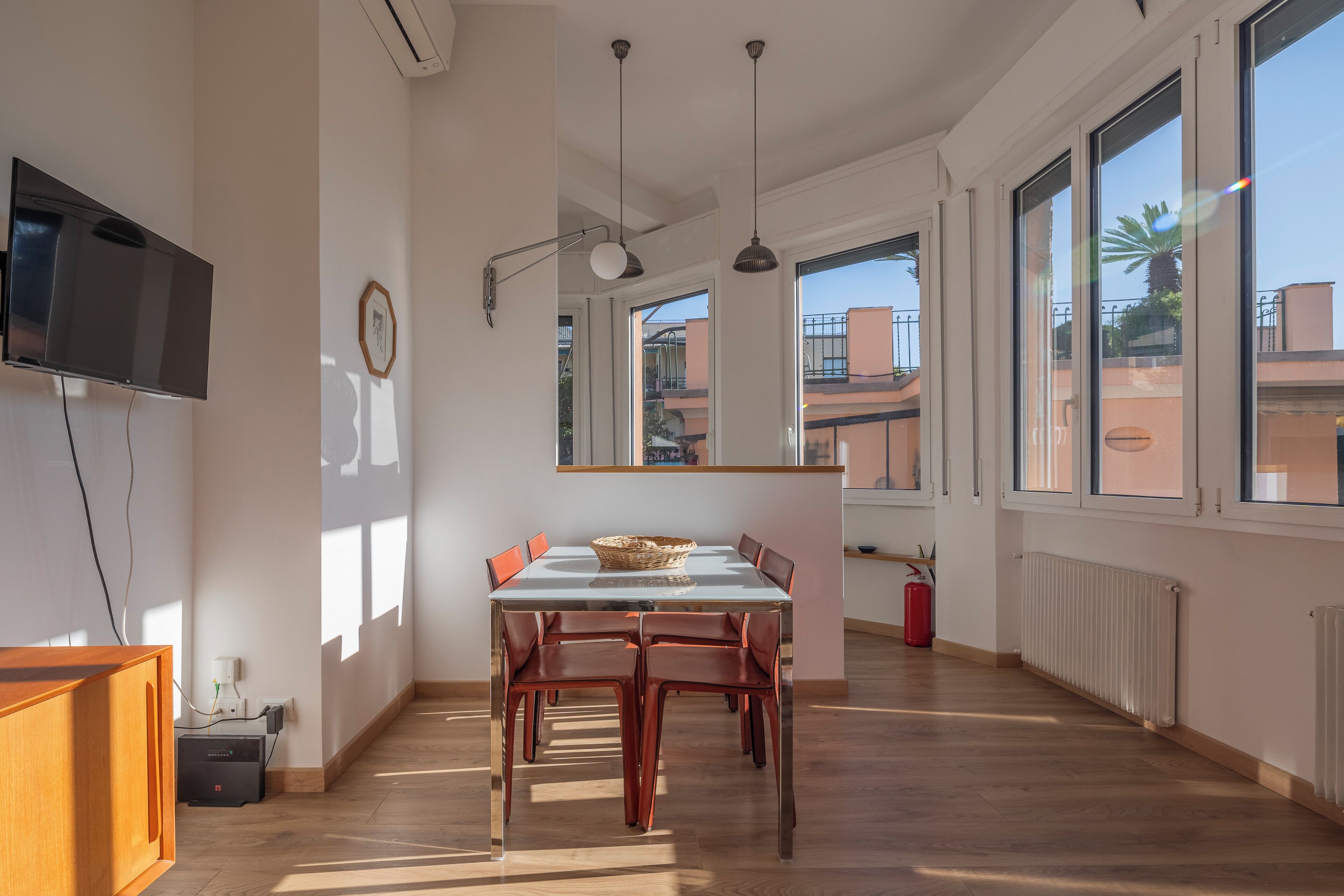 A dining area with a table and chairs, a television mounted on the wall, and large windows with a view of buildings.