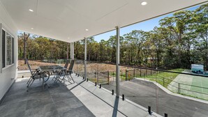 Un espace extérieur abrité, doté d’une table et de chaises, entouré d’une rambarde en verre, et offrant une vue sur un espace paysager avec des arbres et un court de tennis.