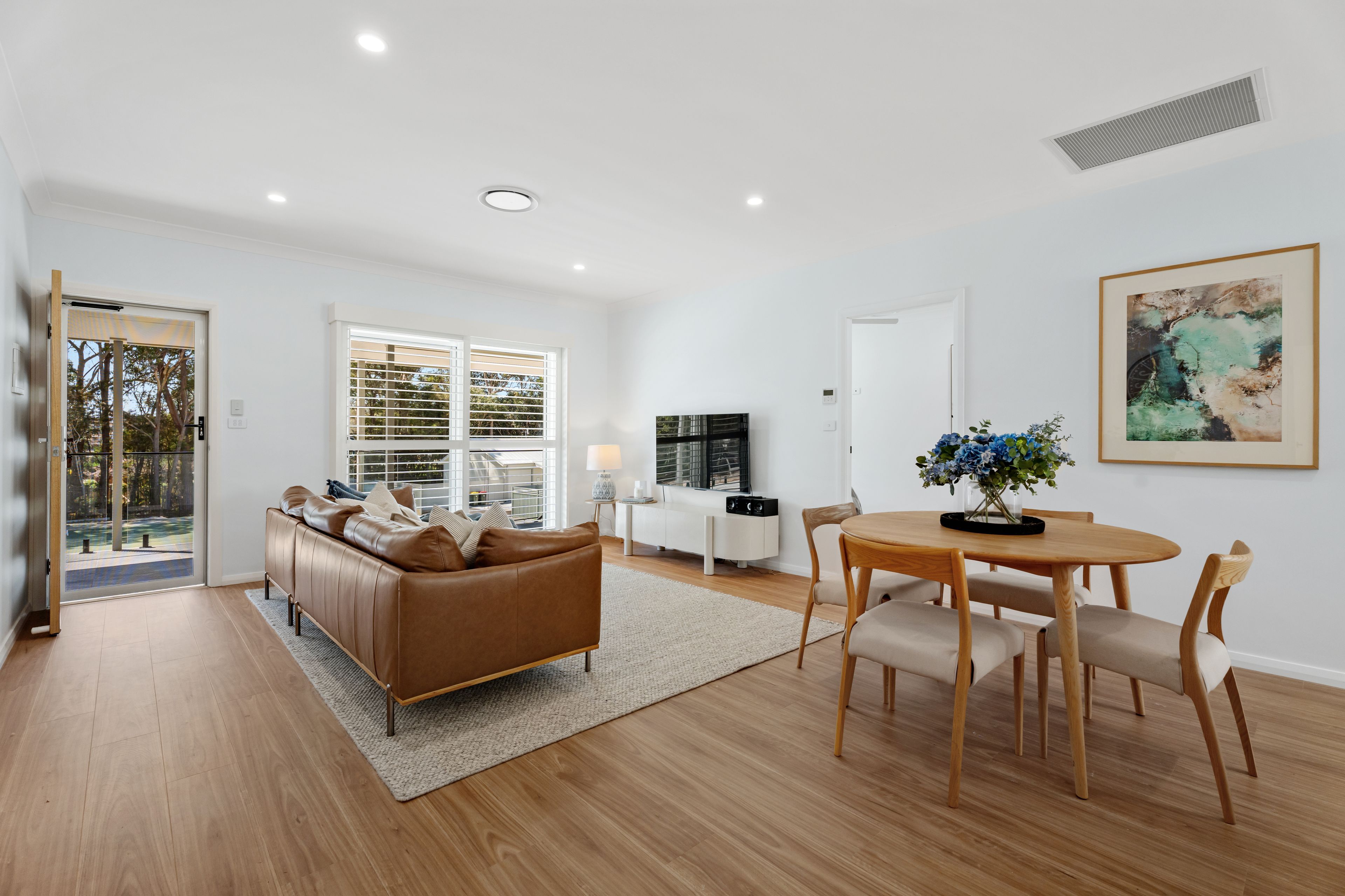 A modern living room with a brown leather sofa, a dining table with chairs, and a painting on the wall.
