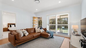 Un salon moderne avec un canapé d’angle en cuir marron, un plancher en bois, un tapis gris et une grande fenêtre avec des stores.