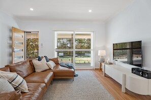 A modern living room with a brown leather sofa, a flat-screen TV, and a large window with blinds.