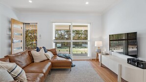 Un salon moderne avec un canapé en cuir marron, un téléviseur posé sur un meuble blanc et une grande fenêtre équipée de stores.