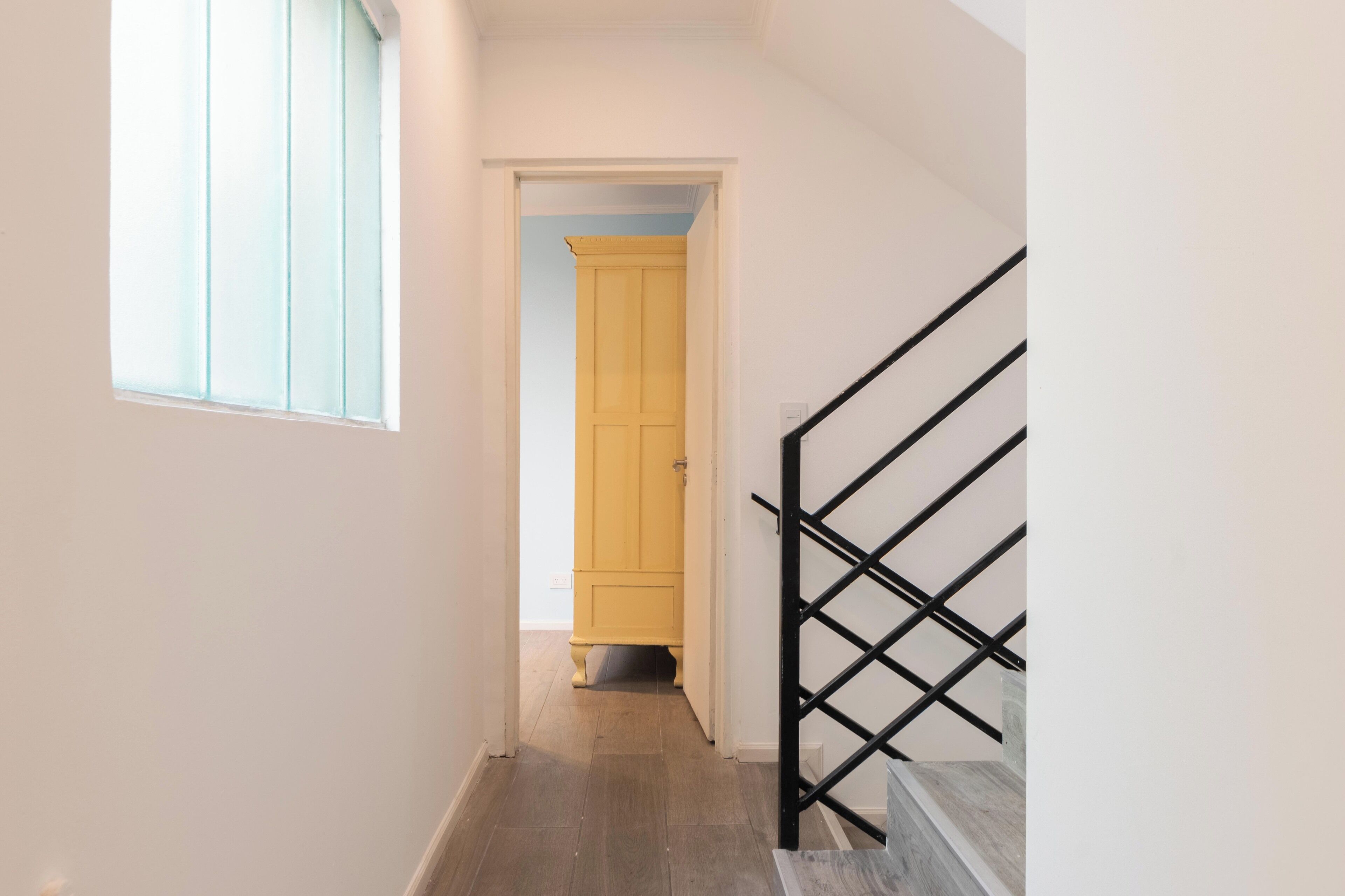 Un couloir étroit avec un plancher en bois, une fenêtre sur la gauche et une porte jaune au fond.