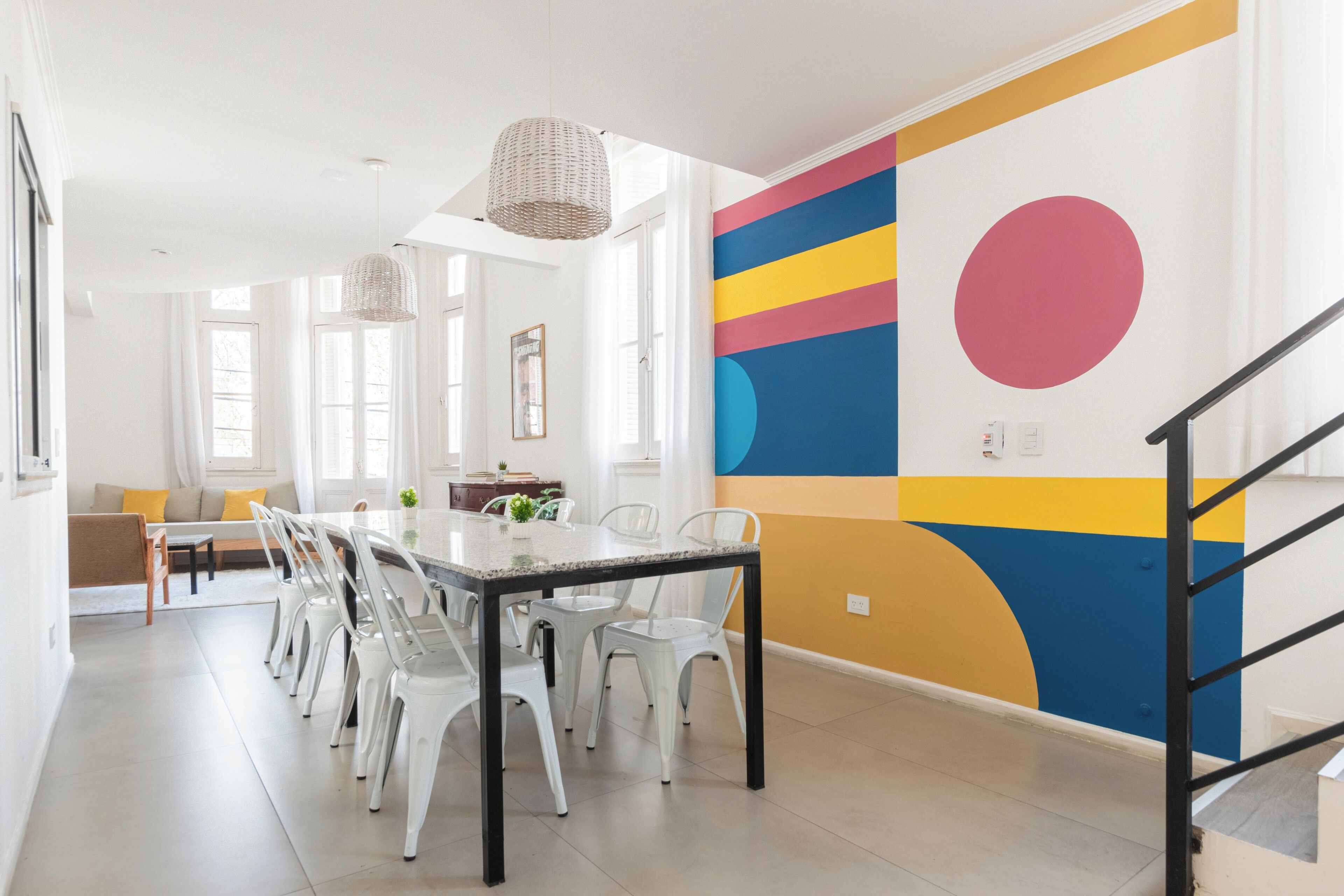 Un comedor moderno con una mesa larga, sillas blancas y un mural de colores en la pared.