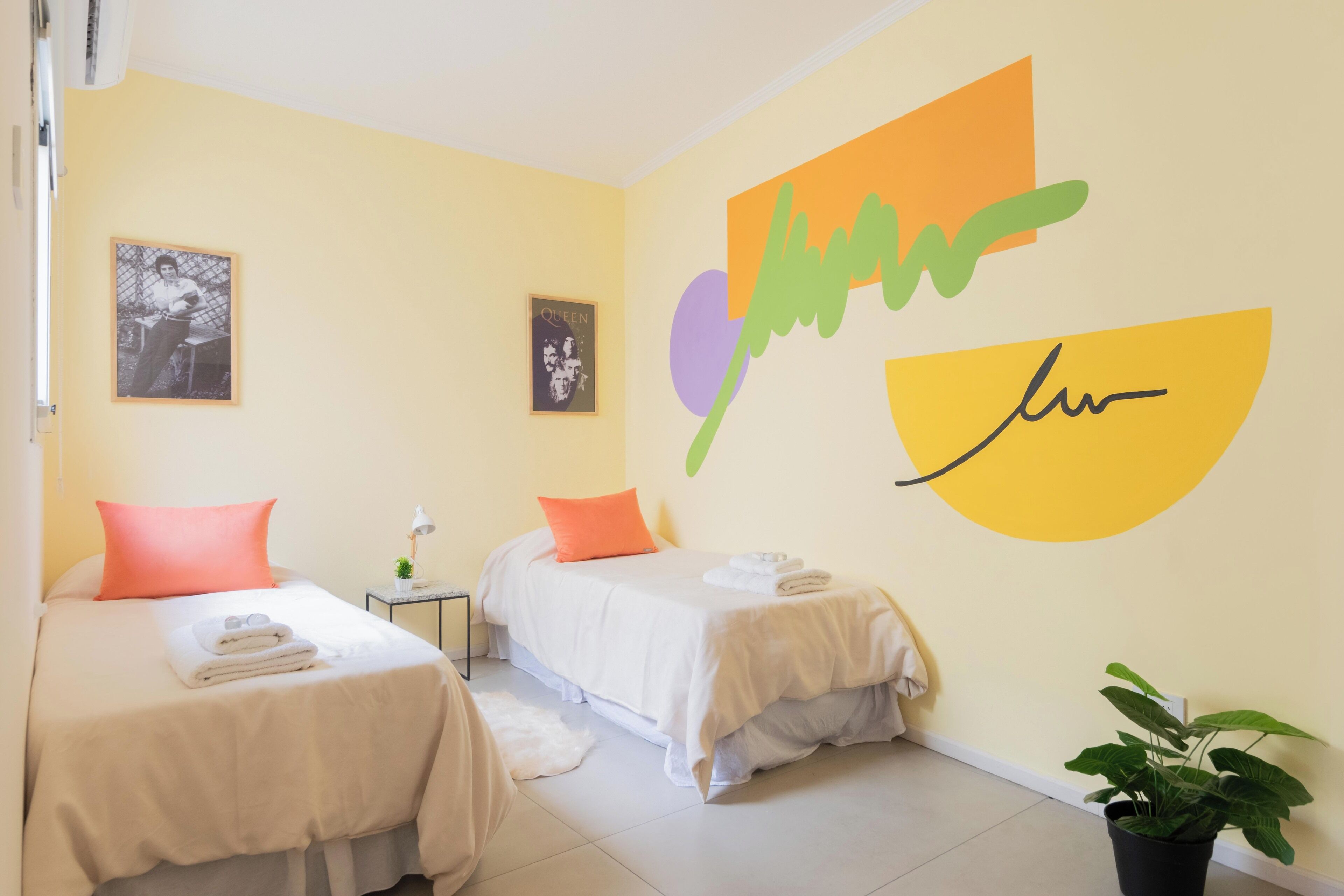 A hotel room with two beds, a small table with a lamp, a potted plant, and wall art.