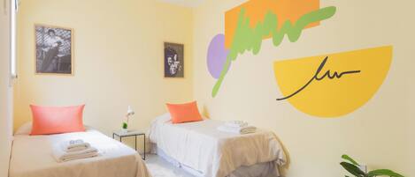 A hotel room with two beds, a small table with a lamp, a potted plant, and wall art.