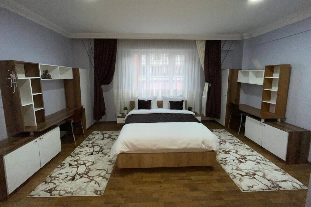 Un dormitorio con una cama centrada, flanqueada por dos armarios con estantes, una ventana con cortinas y una alfombra estampada en el suelo.