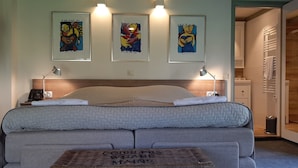Une chambre à coucher avec un lit, des lampes de chevet, une table de chevet, une plante suspendue et trois œuvres d’art encadrées accrochées au mur.