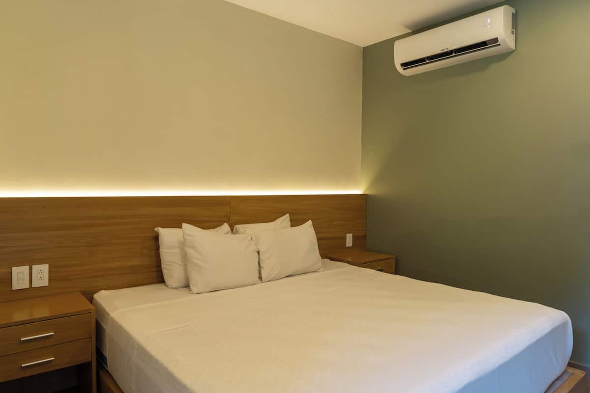 Una habitación de hotel con cabecera de madera, cama con ropa blanca y un equipo de aire acondicionado instalado en la pared.