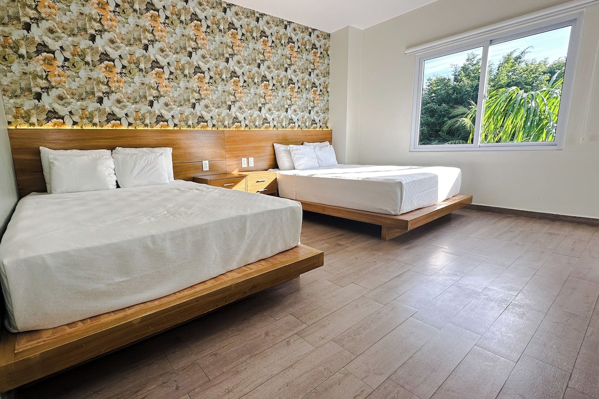 Habitación de hotel con dos camas, cabecero de madera, un ventanal grande y decoración floral en la pared.