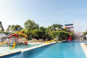 Una piscina con toboganes y zona de juegos acuáticos, rodeada de árboles y un cielo despejado.