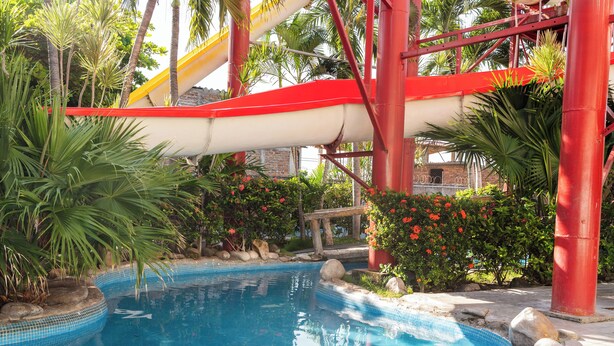 A swimming pool with a water slide, surrounded by tropical plants and rocks. - El Pacifico (La Libertad)