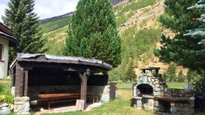 Un espace extérieur rustique aménagé avec des bancs en pierre et une cheminée en pierre, entouré d’un champ herbeux et d’arbres, le tout se fondant dans un paysage montagneux.