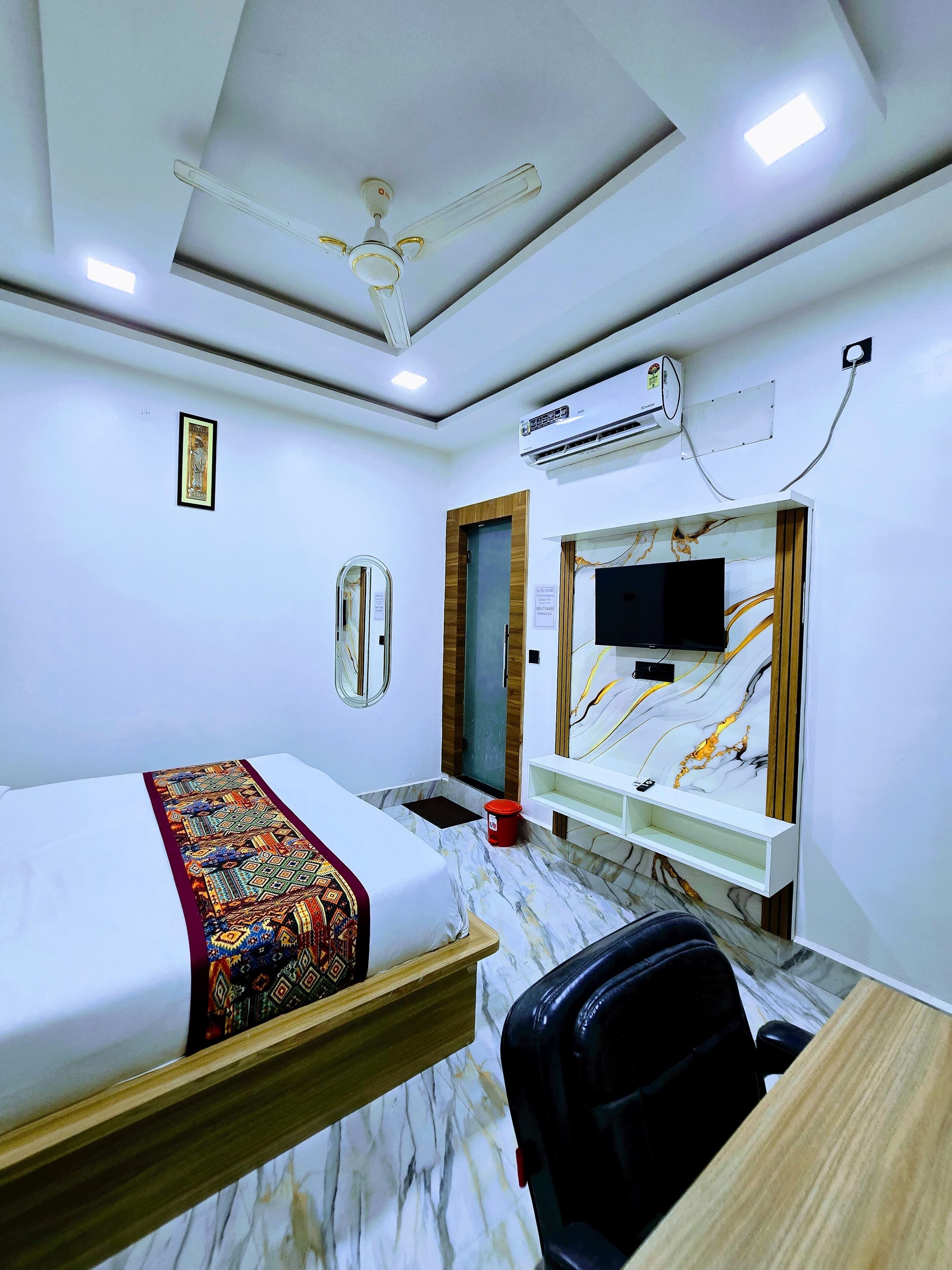 A hotel room with a bed, a desk, a chair, a ceiling fan, an air conditioning unit, and a television.
