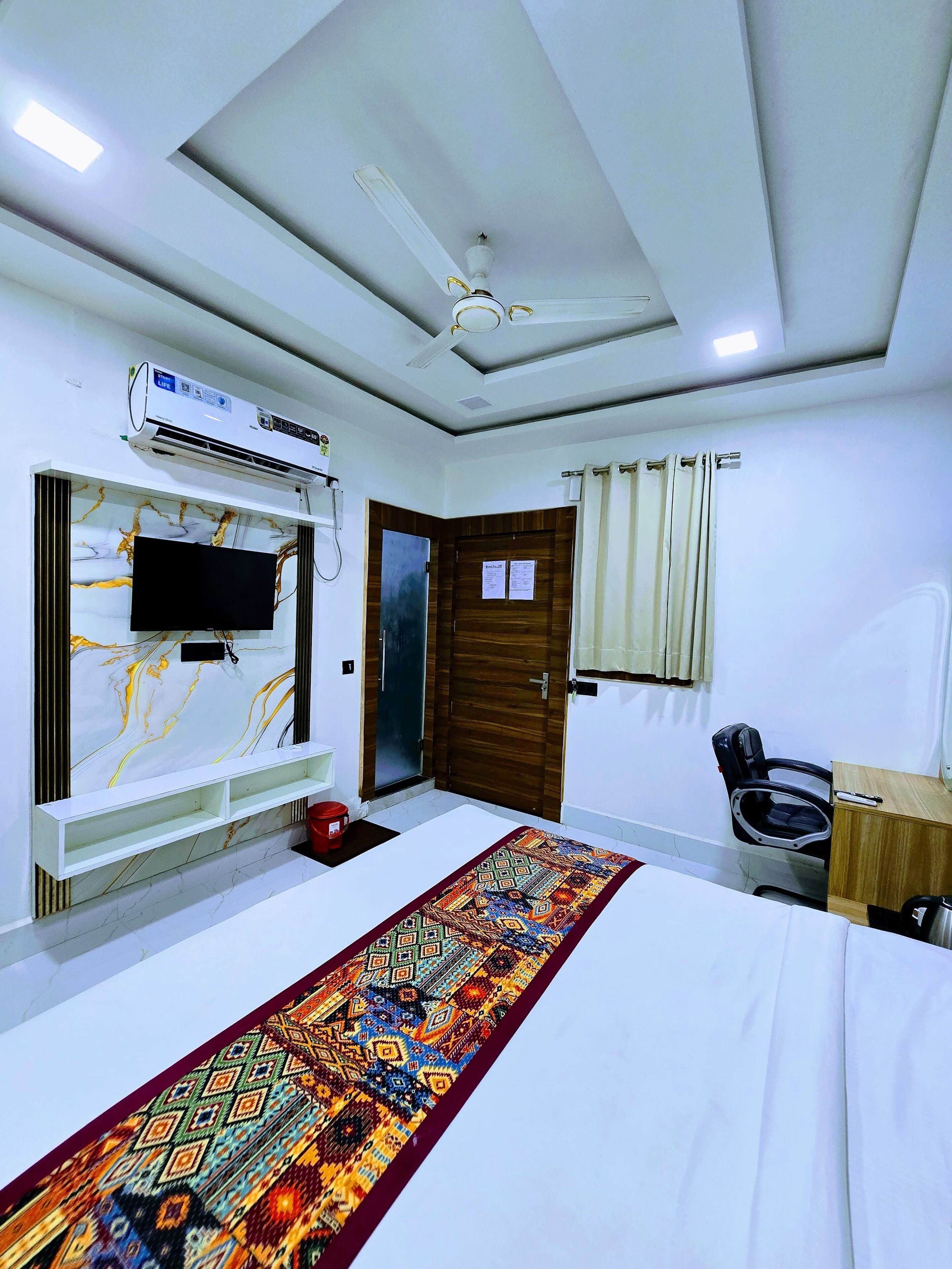A hotel room with a bed, a TV mounted on a marble wall, a ceiling fan, an air conditioning unit, a wooden door, a window with curtains, and a desk with a chair.