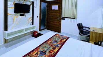 A hotel room with a bed, a TV mounted on a marble wall, a ceiling fan, an air conditioning unit, a wooden door, a window with curtains, and a desk with a chair.