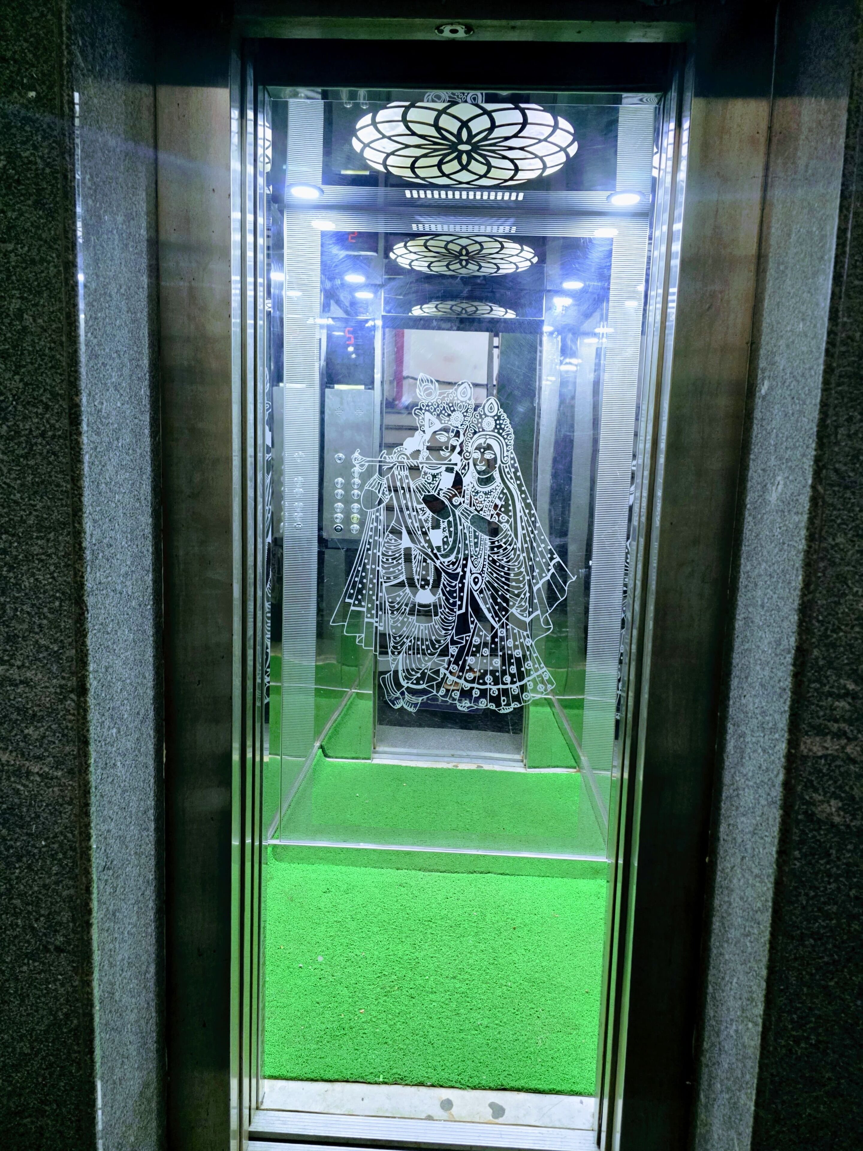 Un ascensor con cabina de vidrio, alfombra verde y una lámpara decorativa en el techo.