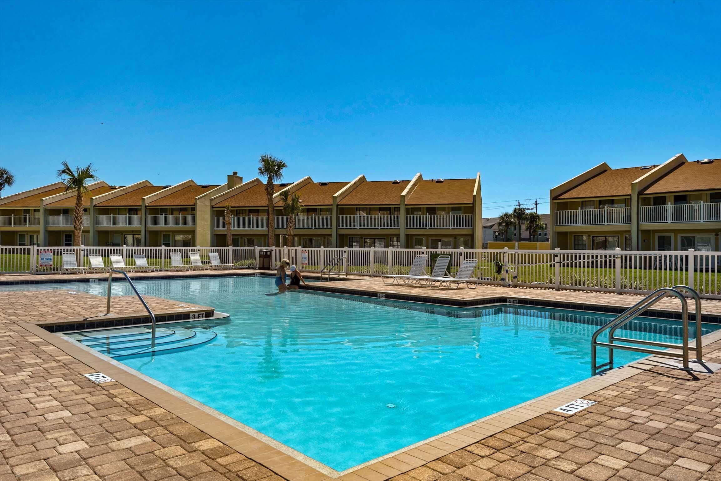 A swimming pool with a tiled deck, surrounded by a white fence and palm trees.