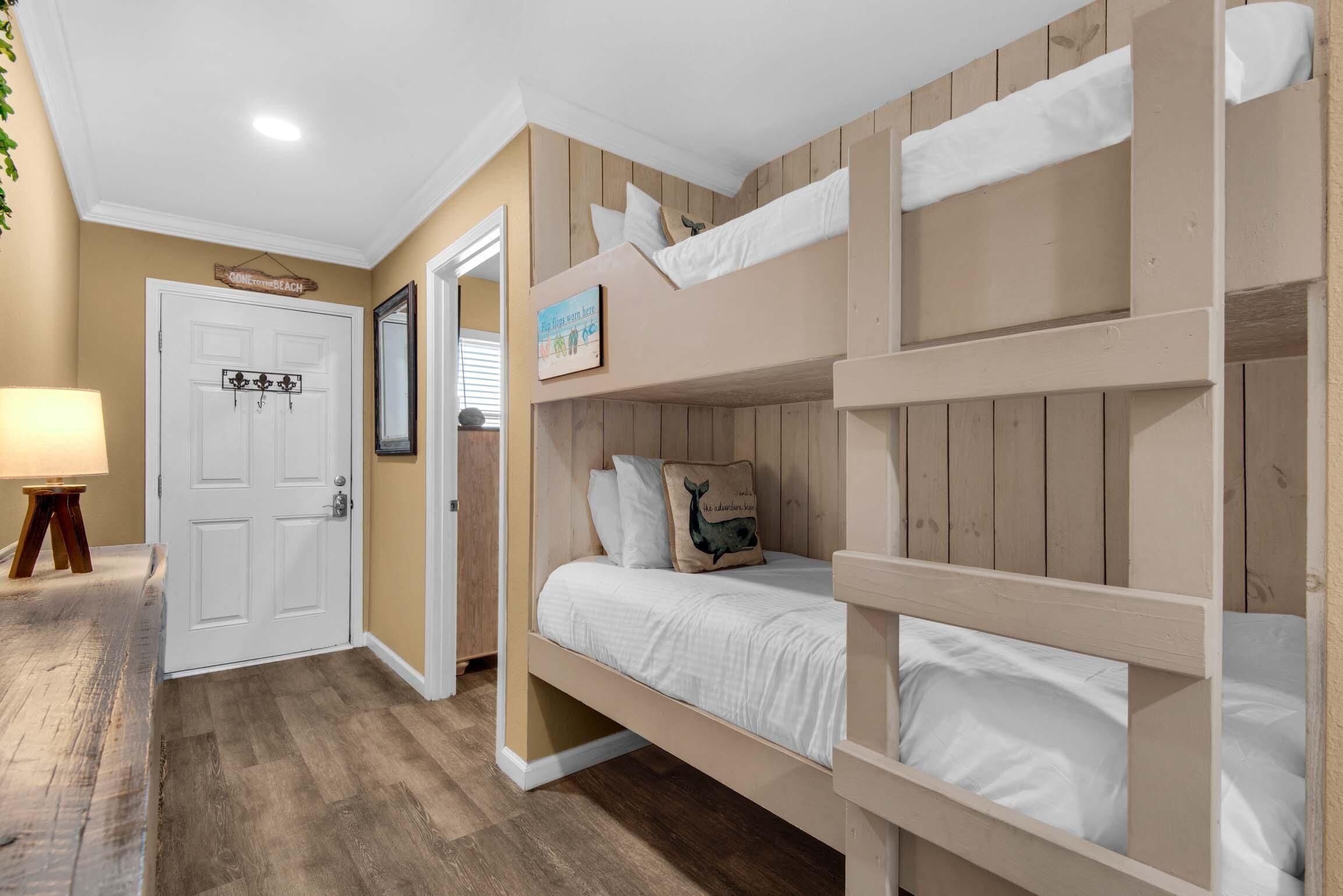 Une chambre avec un lit superposé, un plancher en bois, une porte blanche et une petite table avec une lampe.