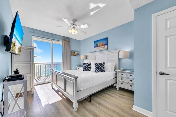 A bedroom with a bed, a nightstand, a TV, a ceiling fan, and a view of the ocean.