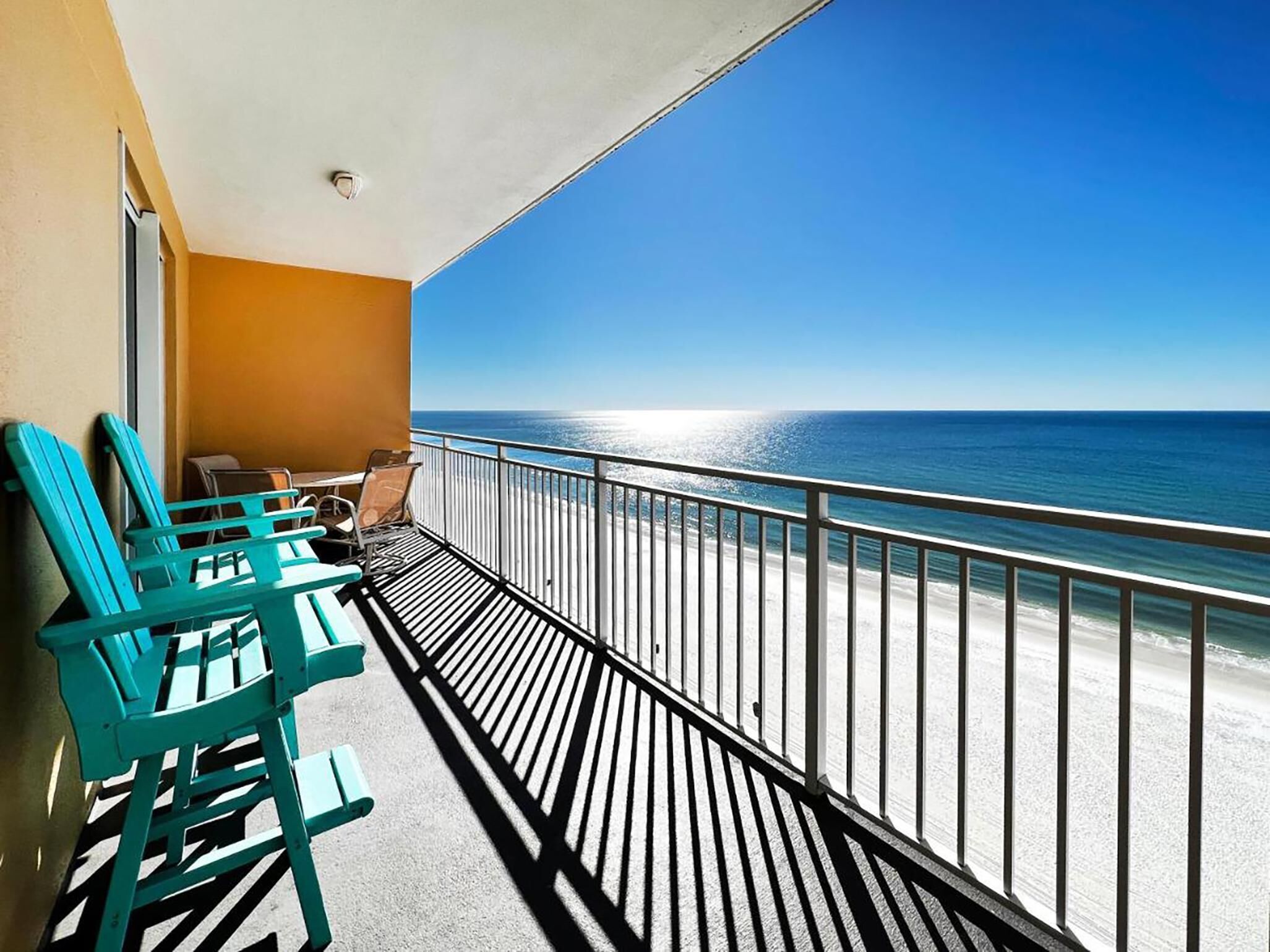 A balcony with turquoise chairs and a table, overlooking a beach with a clear blue sky and ocean.