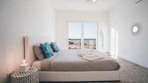 A bedroom with a large bed, a bedside table with a lamp, a view of the sea through a window, and a decorative mirror on the wall. - Villa Bela Vista, beach villa with ocean view  (Atalaia)