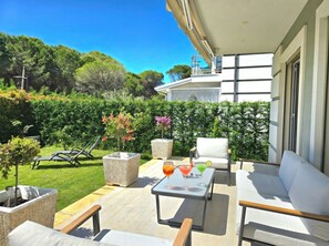 Um pátio com mobiliário externo, uma mesa com bebidas e vista para um jardim exuberante e um edifício.