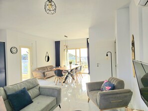 A spacious living area with a sofa, armchair, dining table, and chairs. A large window offers a view of the outdoors.