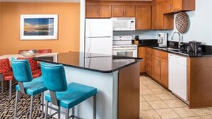 A compact kitchen with wooden cabinets, a white refrigerator, a microwave, and a coffee maker. There are two blue bar stools and a small dining table with a red cushion. - Worldmark Indio Resort/Coachella Week1/2BDR/Sleeps 6 (Indio)