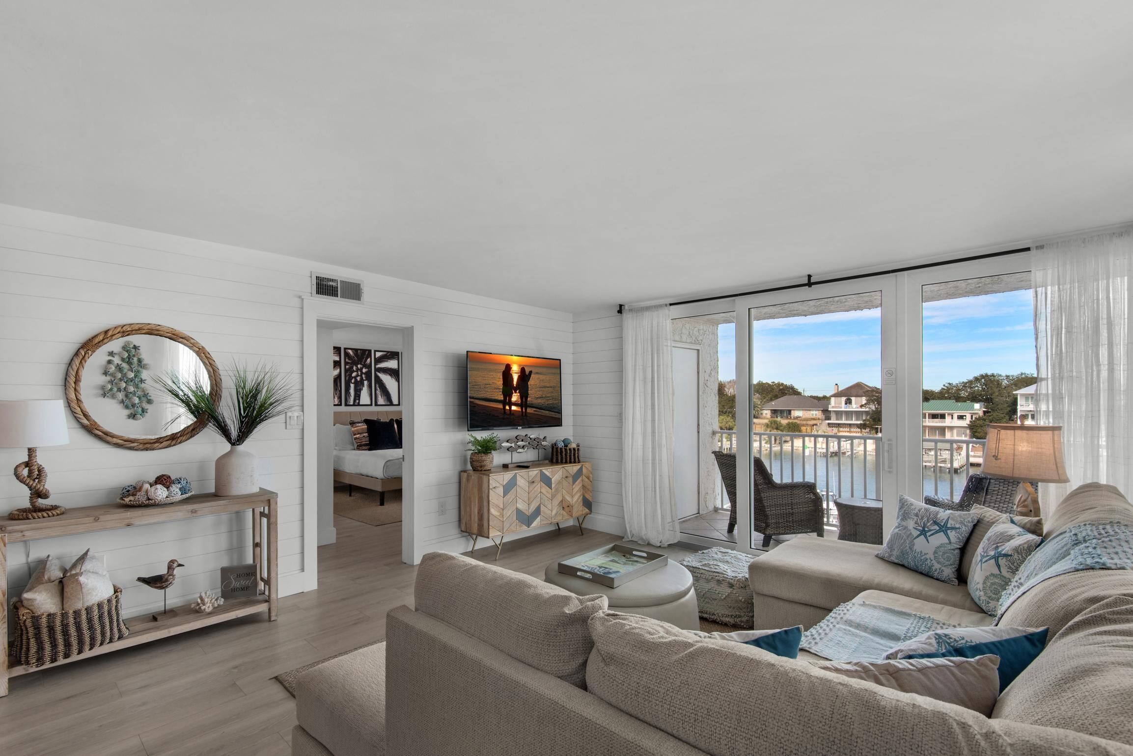 A spacious living room with a large window offering a view of a body of water and buildings. There is a comfortable sofa, a wooden console table with decorative items, and a flat-screen TV mounted on the wall.