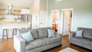 A modern living room with two grey sofas, wooden bar stools, and a kitchen area with a stainless steel range hood. - Charming 2-bedroom house in Wonderful Bolivar Peninsula with AC, WiFi (Bolivar Peninsula)