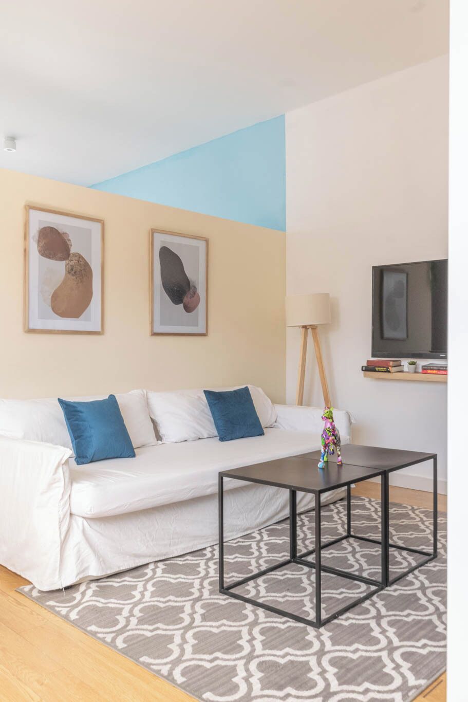 A modern living room with a white sofa, two framed abstract paintings, a black coffee table, a lamp, and a television on a stand.