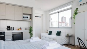 Ett modernt hotellrum med en säng, ett litet pentry, ett fönster med utsikt över staden och ett nattduksbord med en växt.