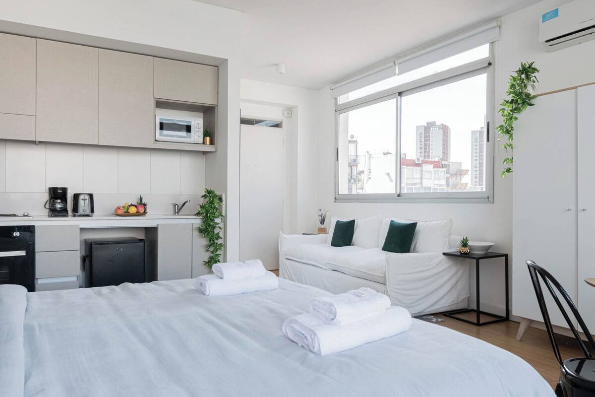 Una habitación de hotel moderna con cama, una kitchenette pequeña, un ventanal con vista a la ciudad y una mesita de noche con una planta.