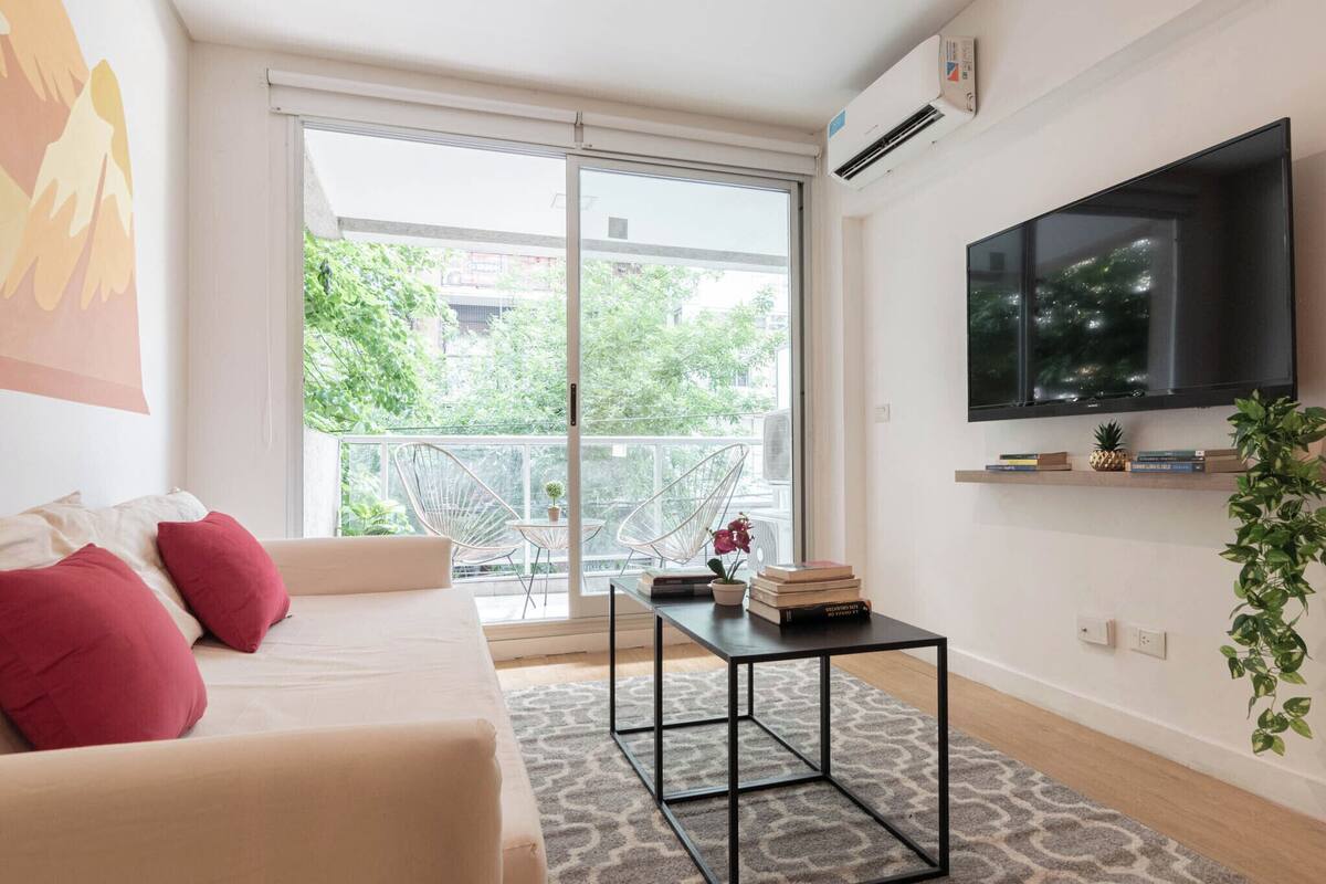 Una sala de estar moderna con un sofá, una mesa de centro, un televisor montado en la pared y un balcón con vistas a zonas verdes.