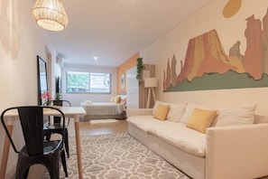 A room with a white sofa, a dining table with chairs, a lamp, and a painting of mountains.