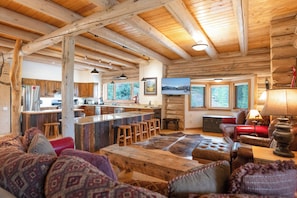 A rustic cabin interior with a wooden ceiling, a kitchen area, a bar, and a seating area with a large rug.