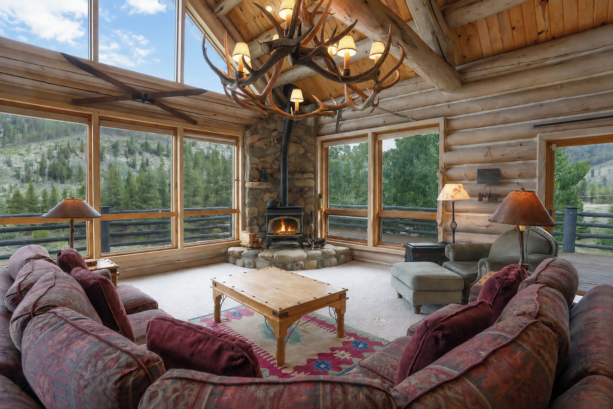 A log cabin interior with a stone fireplace, large windows, and a ceiling chandelier.