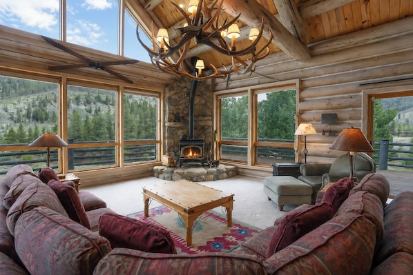 A log cabin interior with a stone fireplace, large windows, and a ceiling chandelier.