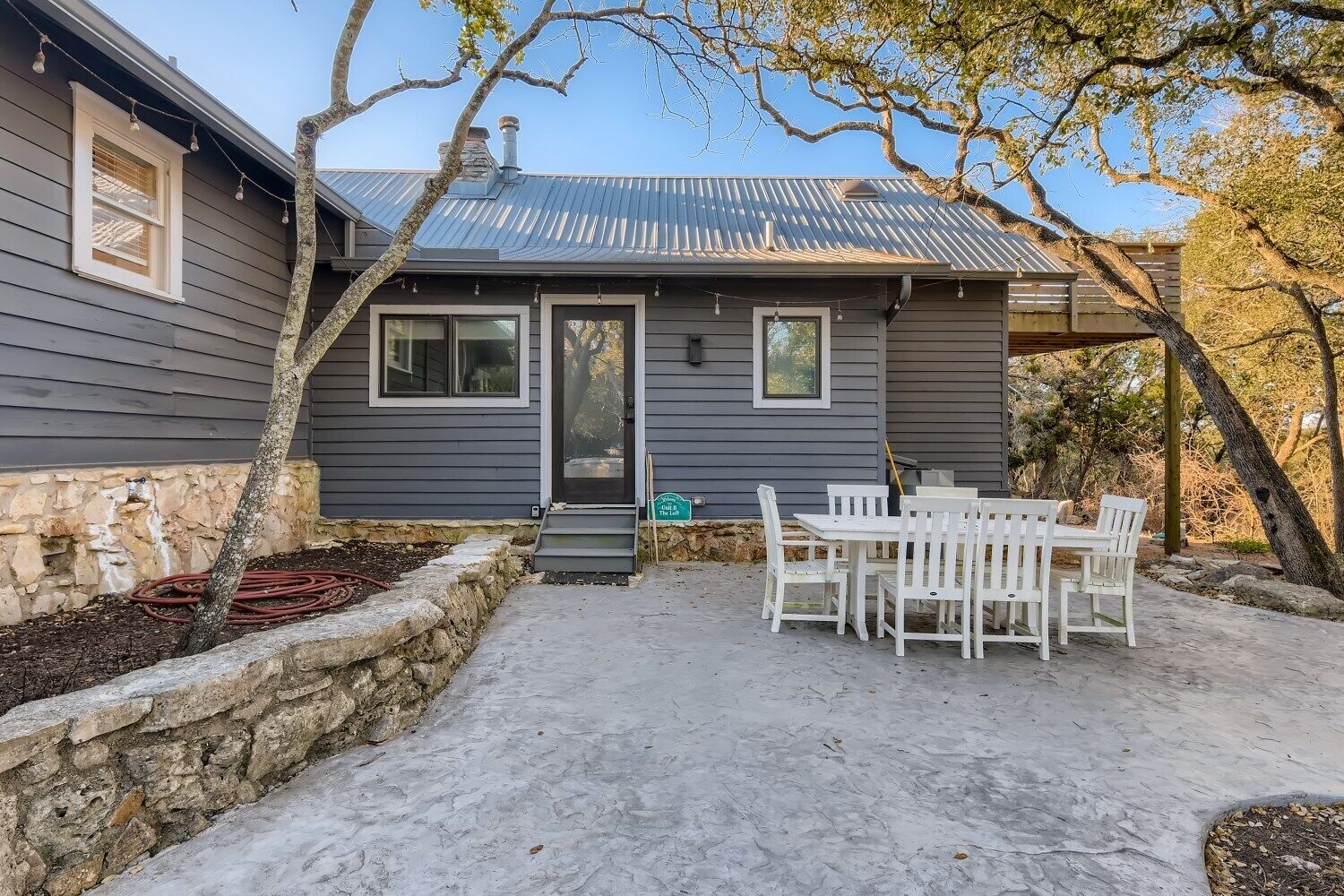 Une maison avec un mur de soutènement en pierre, un toit en métal et une terrasse avec une table et des chaises blanches.